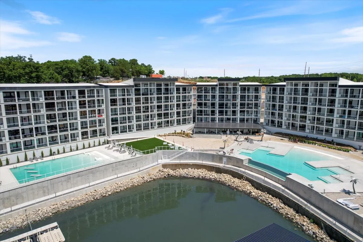 This image shows a vibrant community pool area set against modern architecture and a waterfront view. The area is bustling with activity, featuring people swimming, sunbathing, and socializing in well-landscaped gardens. The setting radiates a resort-like ambiance, with the pool as a lively focal point for community events and outdoor dining.