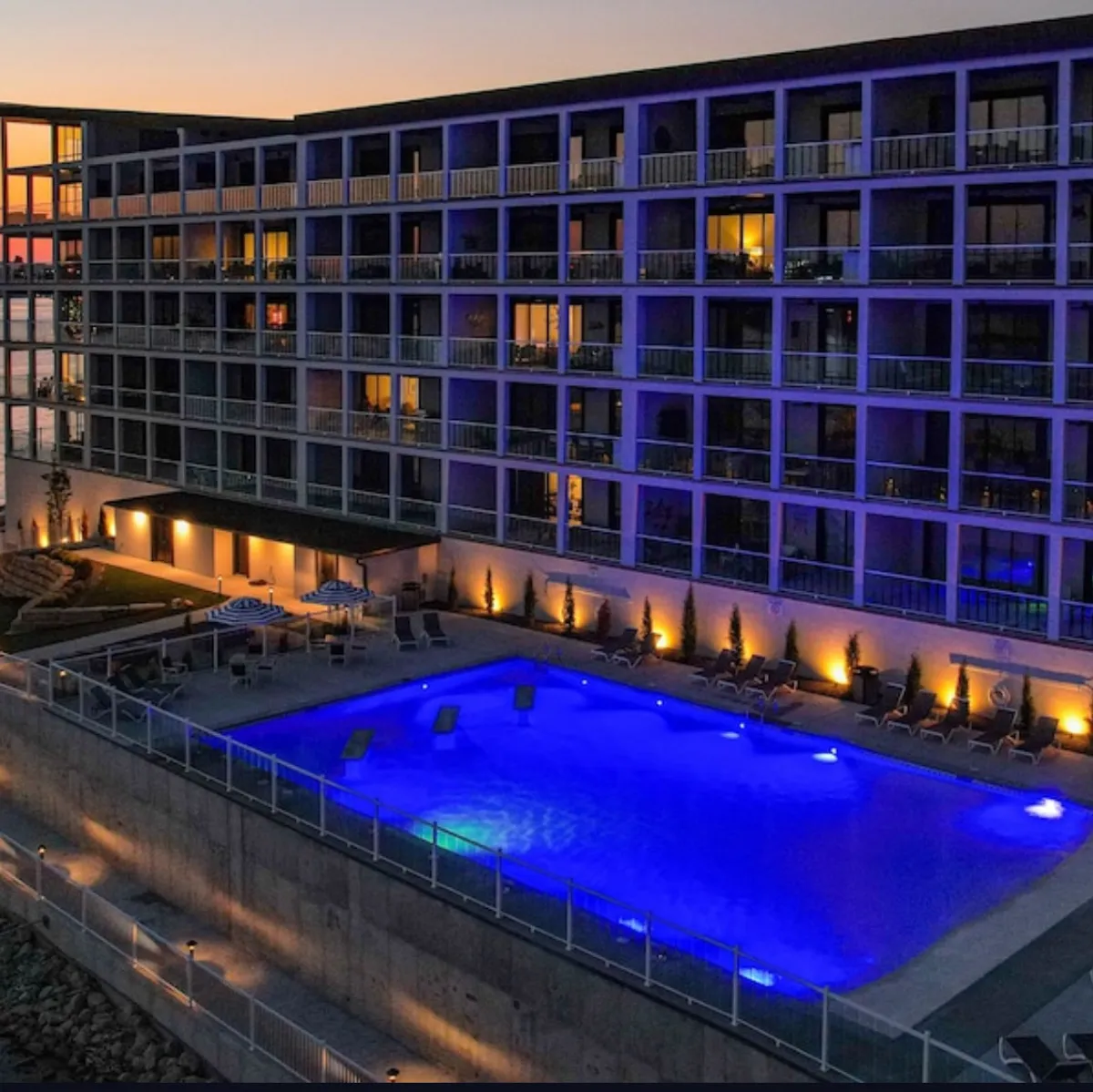 Taken at dusk, this image shows an illuminated pool with warm glowing balconies above and a landscaped area with comfortable seating. The tranquil, inviting ambiance suggests evening relaxation, stargazing, and family gatherings around the pool after a day of activity.