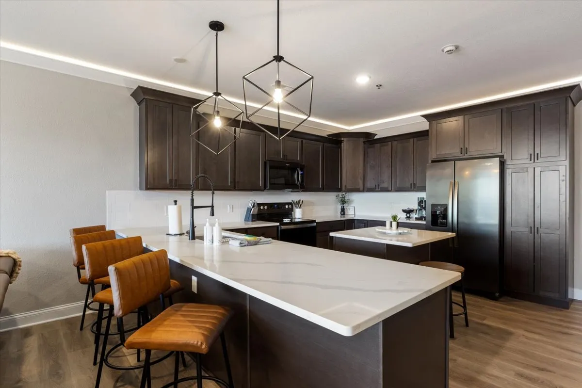 A bright, welcoming kitchen area with a granite island, stylish hanging light fixtures, and bar stools. The scene evokes gatherings around the island—whether for morning coffee, casual meals, or evening cocktails—with a sense of contemporary comfort and connection.
