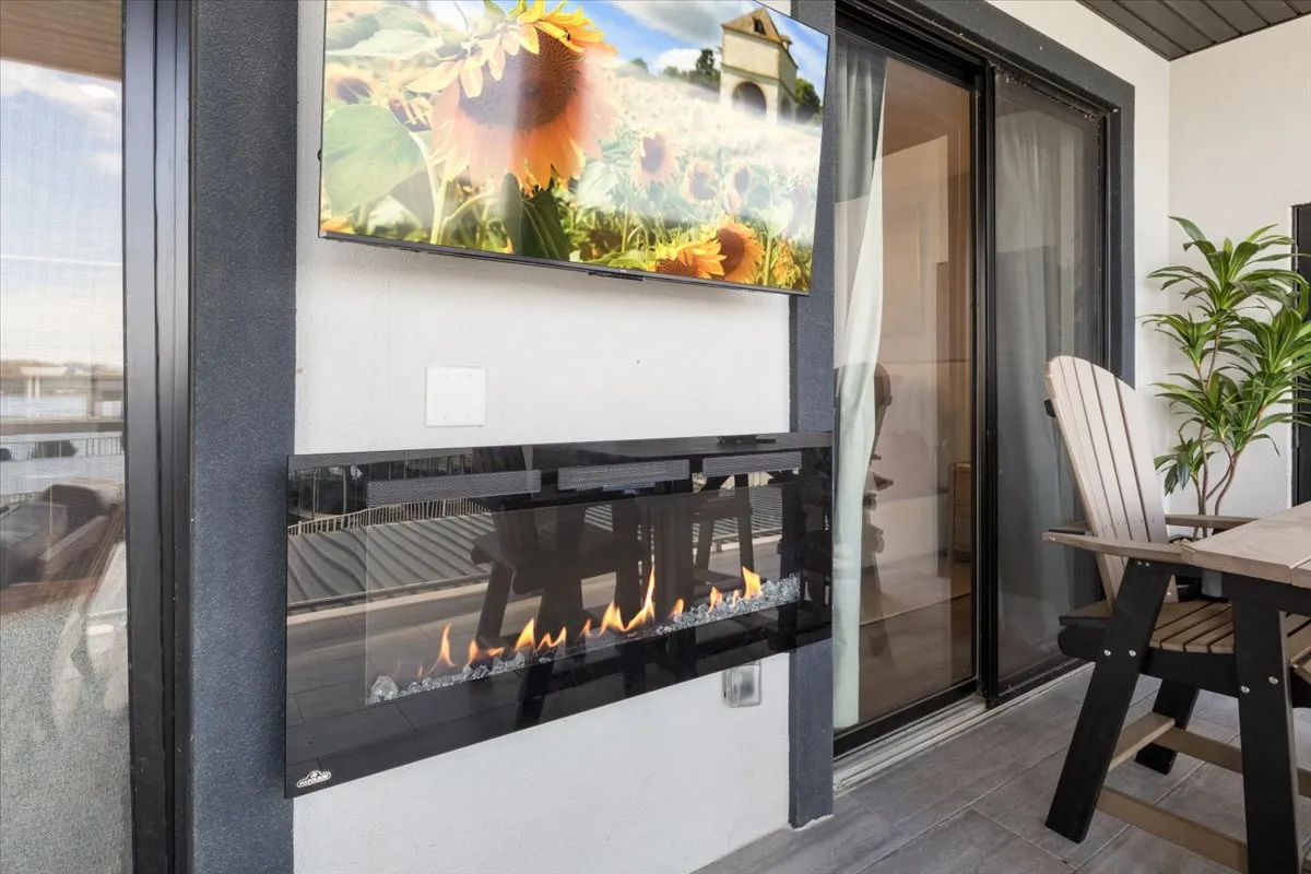 A modern outdoor living area with a sleek fireplace, wall-mounted television, dining table, and lush green plants. The setup is designed for comfort and versatility, making it suitable for both casual lounging and lively gatherings. Warm light and natural elements create a cozy, inviting ambiance.