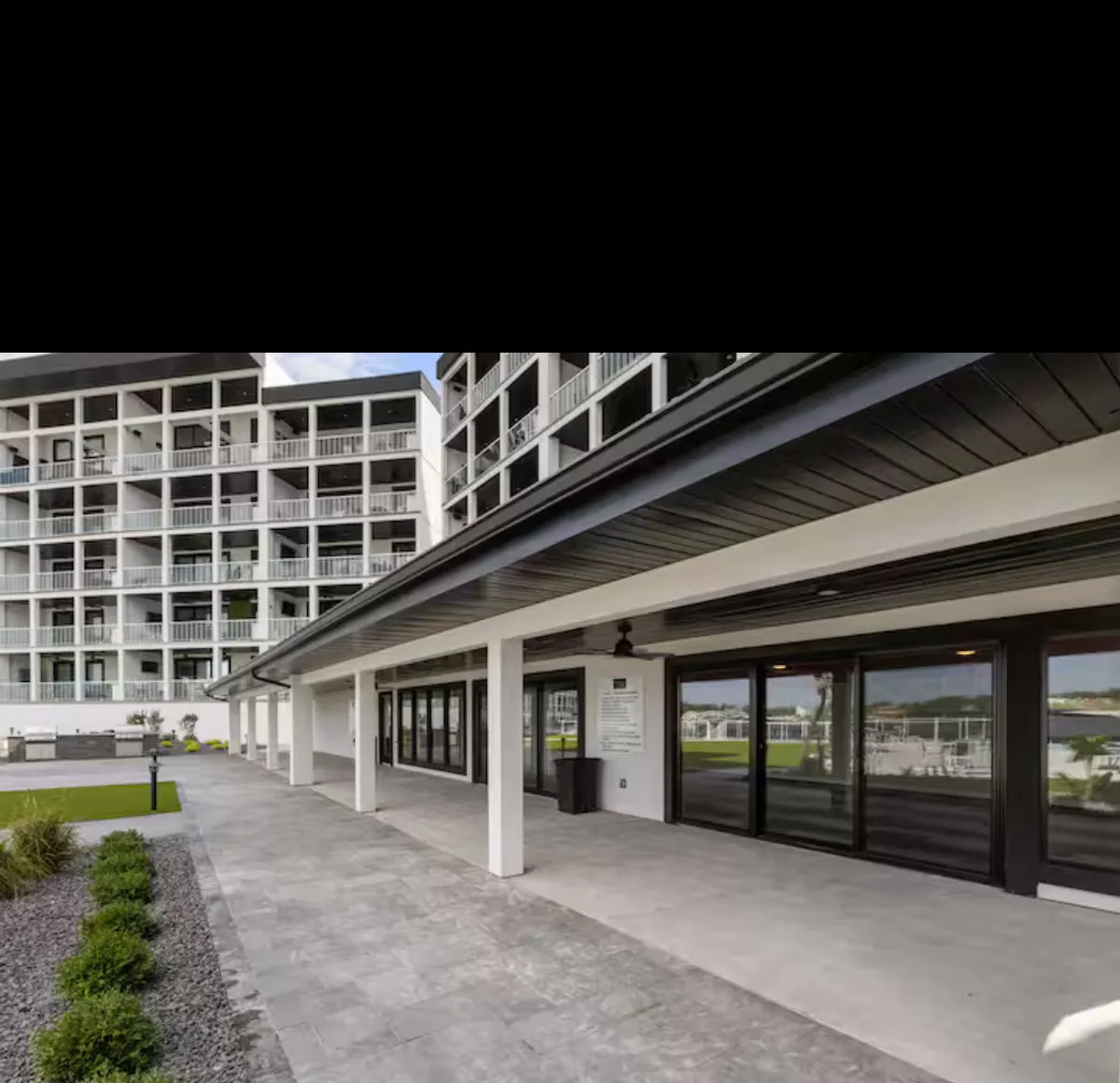 This exterior image features the balconies of the building, a paved walkway, and landscaped greenery in the foreground. The scene evokes a modern, community-oriented atmosphere, with visible outdoor spaces ideal for strolls or casual socializing.