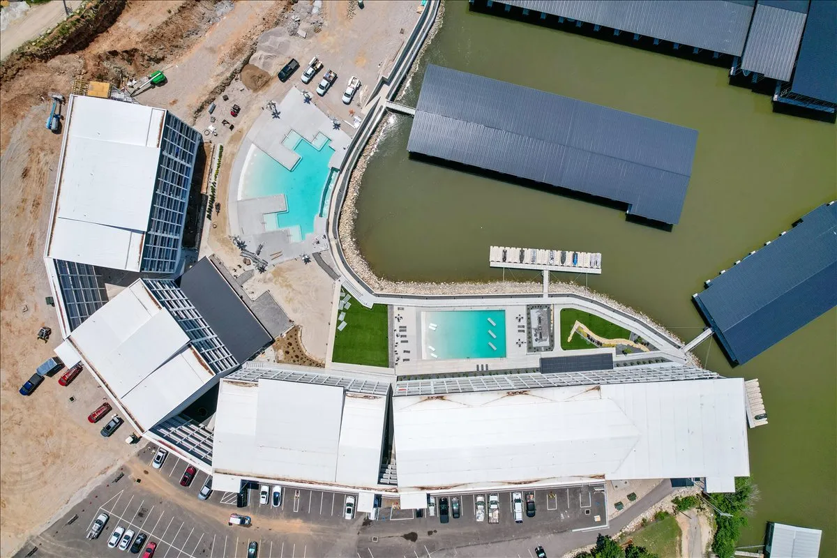 This image combines a vibrant swimming pool area, marina, and landscaped green space, all set against modern buildings. The marina is visible beyond the pool, and the scene is lively yet serene, with places for relaxation and social gathering.