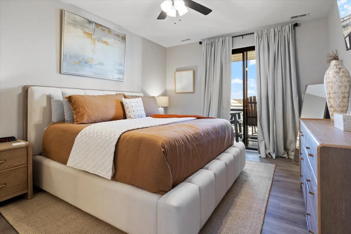 A spacious bedroom featuring a large bed with rich brown bedding, stylish artwork above the headboard, and a balcony door that lets in abundant natural light. The room exudes calm and comfort, perfect for unwinding after a day by the lake.