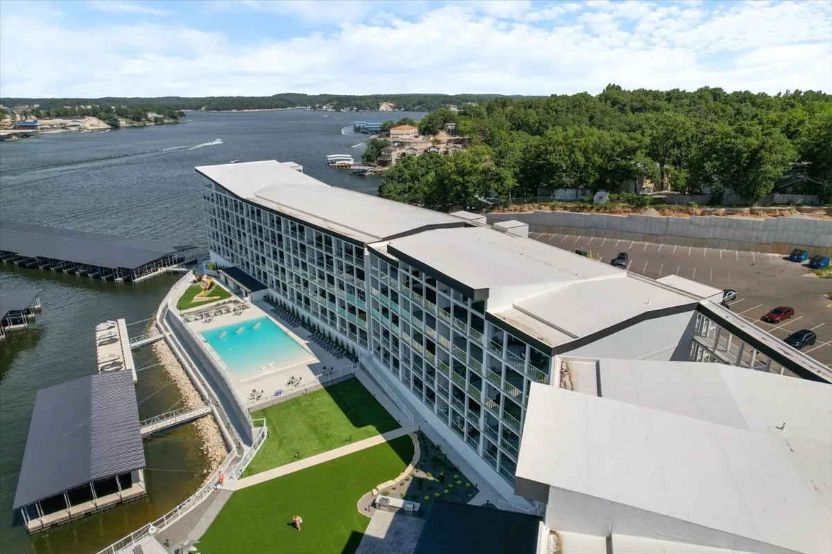 This image displays a sunny exterior view featuring a sparkling swimming pool, a well-kept lawn area perfect for children to play, a waterfront in the background, and the stylish facade of the resort buildings. The combination of the pool, open green space, and proximity to the water captures the essence of outdoor family fun and lakeside leisure.
