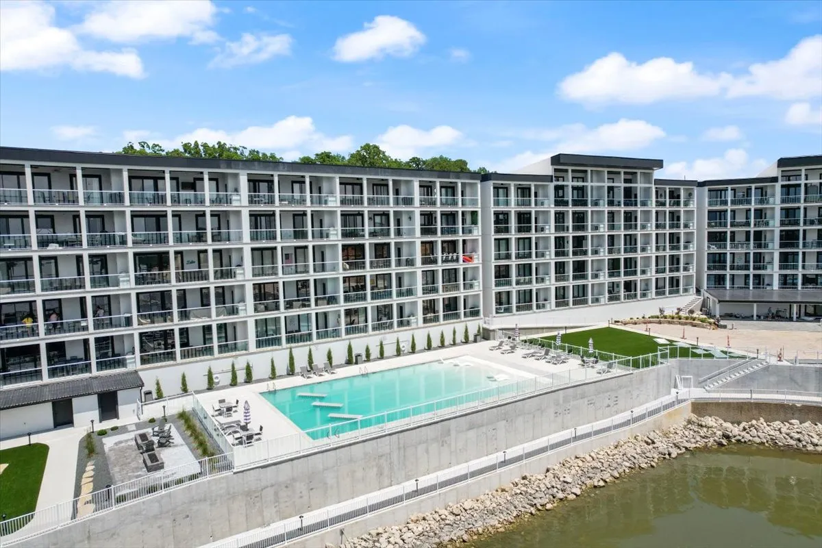 A sleek, modern building faces onto a shimmering swimming pool, surrounded by lush landscaping and a tranquil waterfront. Poolside lounge chairs invite relaxation, while balconies with seating hint at private moments overlooking the water. The space is bathed in golden sunset light, creating an atmosphere of luxury and community.