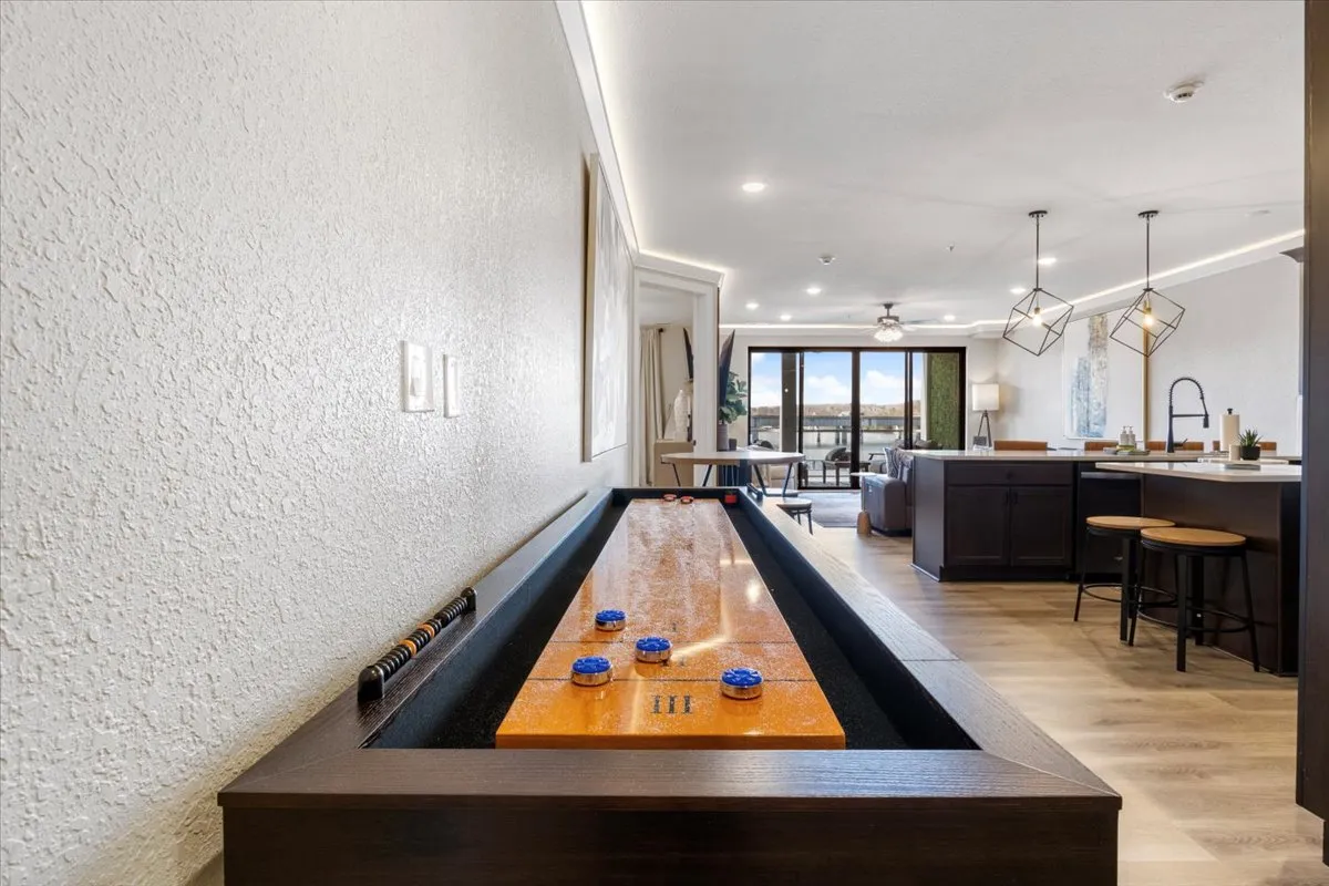 This image presents the open concept living area, prominently featuring the in-unit shuffleboard table, large windows with stunning lake views, and cozy, contemporary furnishings. The scene is lively and playful, inviting guests to relax or engage in friendly competition while basking in natural light.