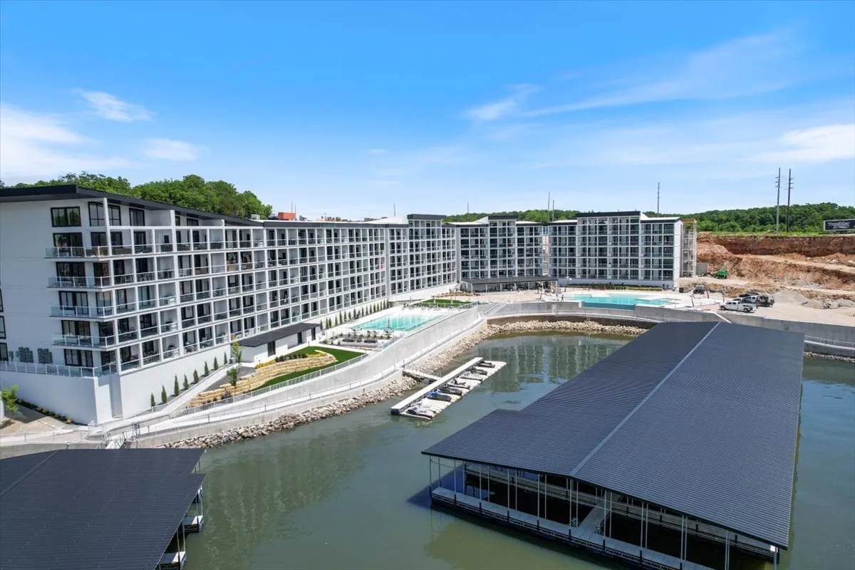 This exterior image captures a modern apartment building with a swimming pool in the foreground and a marina with boats in the background. The pool area is inviting with sun loungers, while the marina signals direct access to lake adventures and boating activities. The setting sun casts a relaxed, vibrant mood over the entire scene.