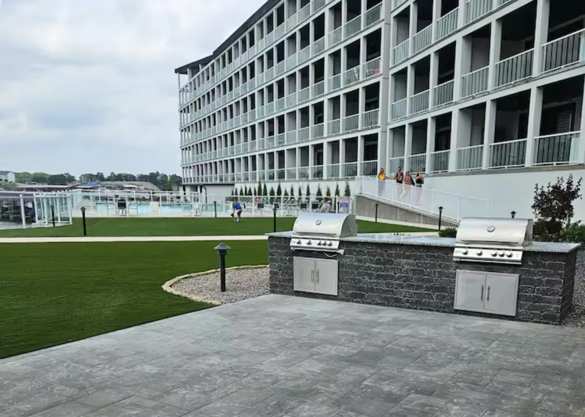 The image captures a welcoming BBQ area adjacent to a green lawn and the pool. The scene includes a grilling station, outdoor dining area, and guests gathering for a casual cookout, with the pool and building exterior in the background. The atmosphere is inviting and ideal for social gatherings.